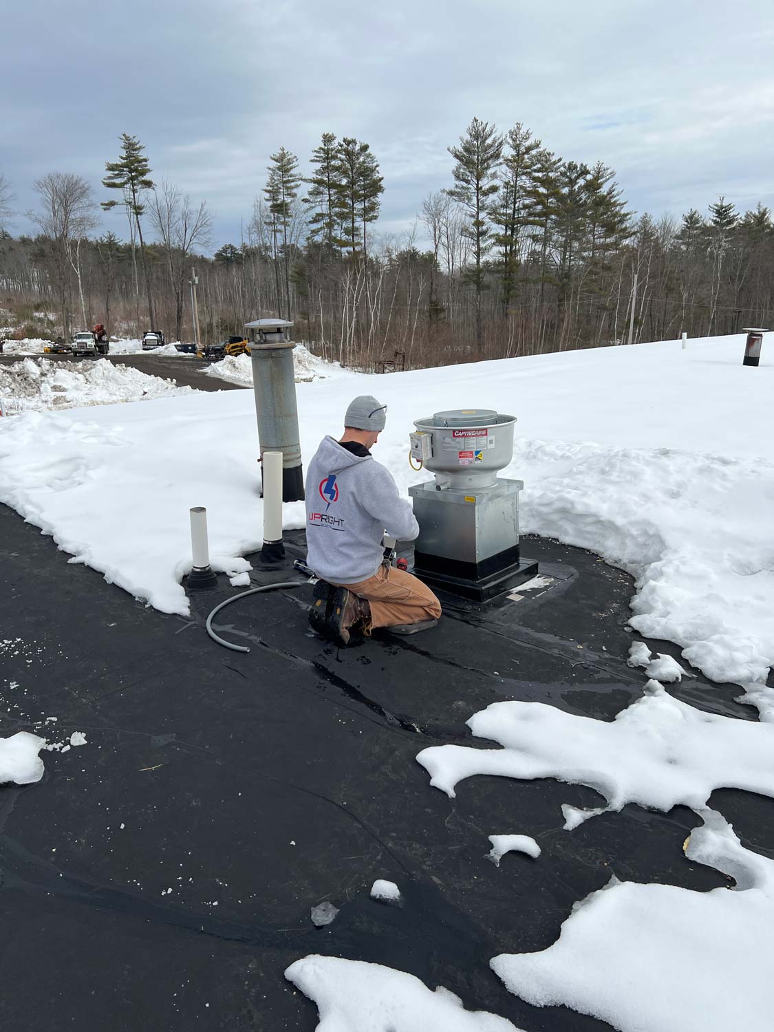 Upright Electric technician wiring a rooftop CaptiveAire exhaust fan on a snow-covered commercial roof