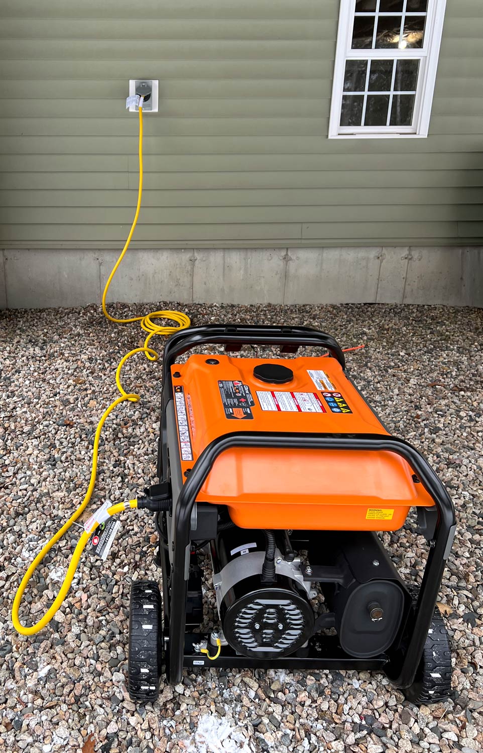 Portable generator on gravel with a yellow cord plugged into a weatherproof inlet on a Lakes Region home