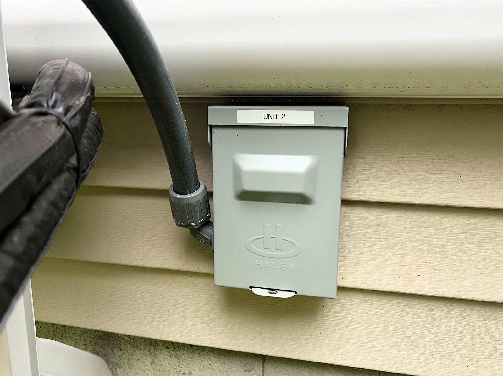 Weatherproof disconnect labeled Unit 2 with a flexible conduit whip on the siding beside a mini-split condenser