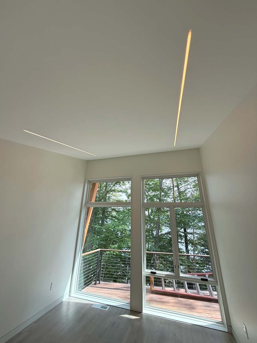 Two recessed linear ceiling lights above hardwood floors and tall windows looking onto a wooded deck