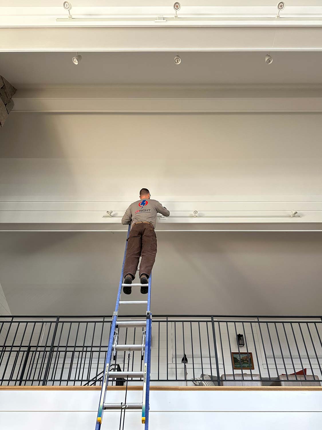 Upright Electric technician on an extension ladder installing track lighting on a high residential wall